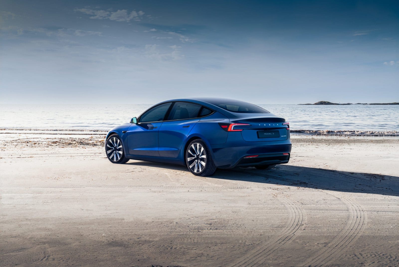 A blue Tesla Model 3 electric vehicle parked on a sandy beach with the ocean in the background under a clear sky.