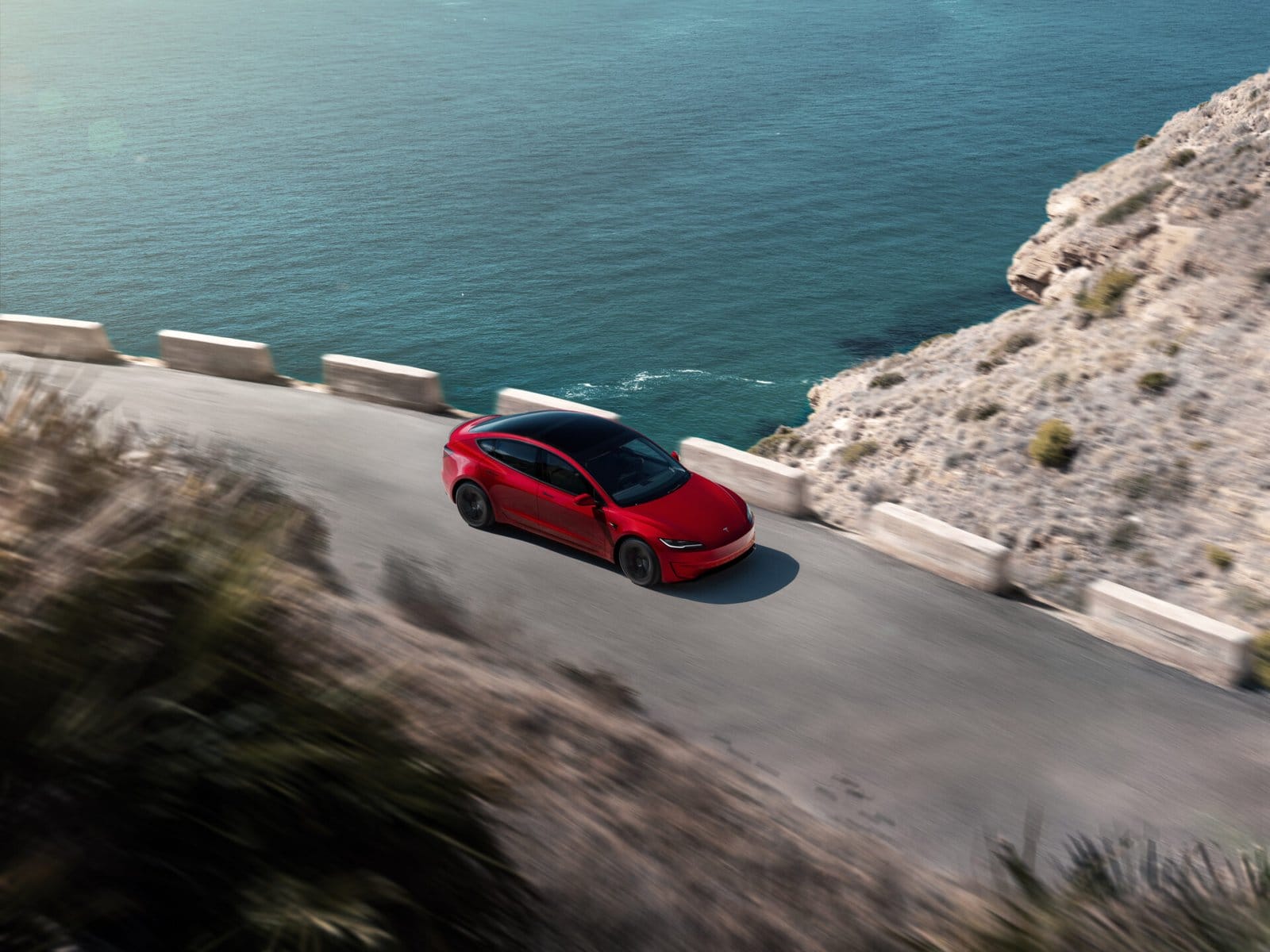 A red Tesla Model 3 electric vehicle navigating a winding coastal road with an ocean view in the background.