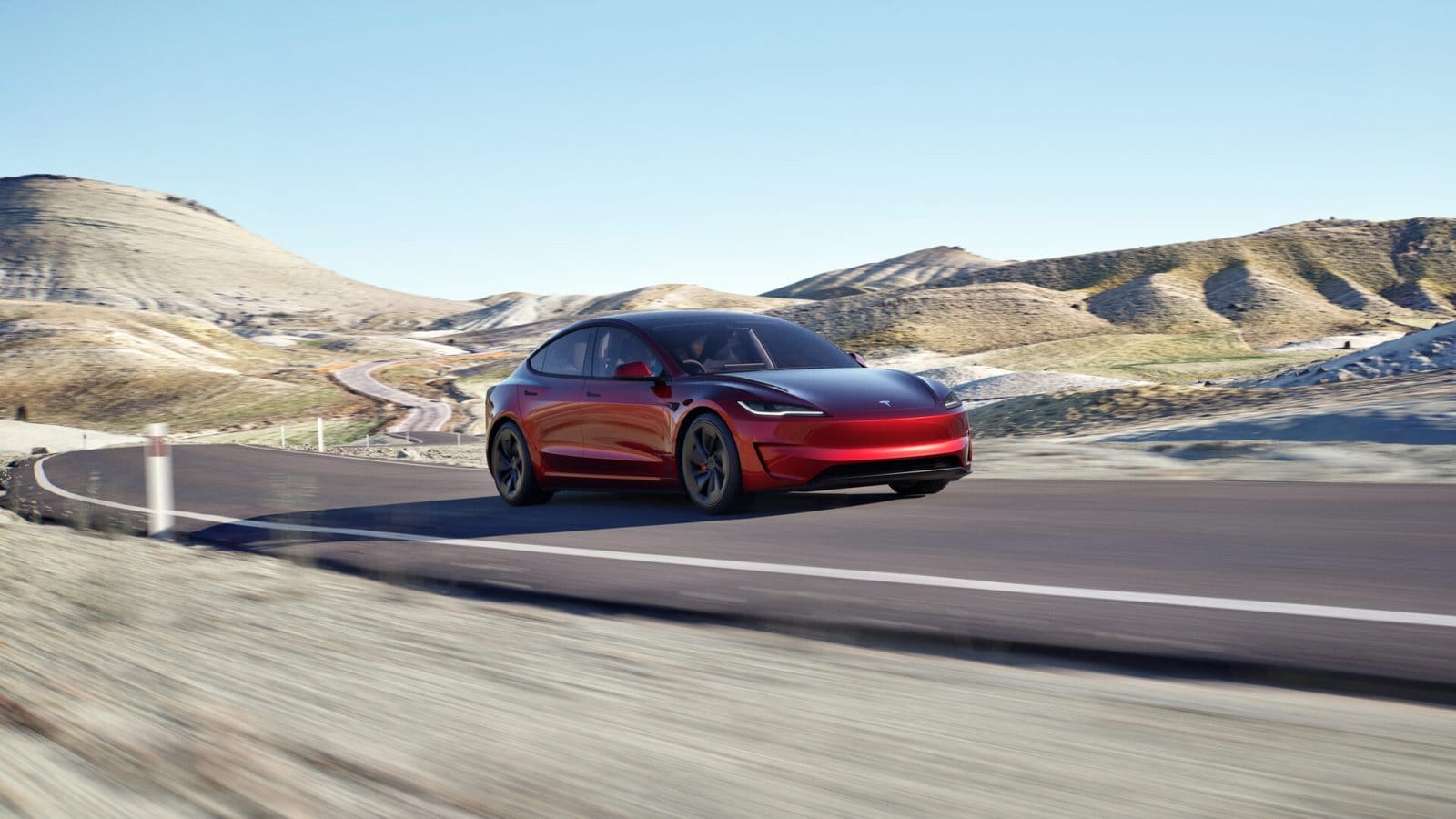 A red Tesla Model 3 electric vehicle driving along a winding road in a mountainous desert landscape under a clear blue sky.