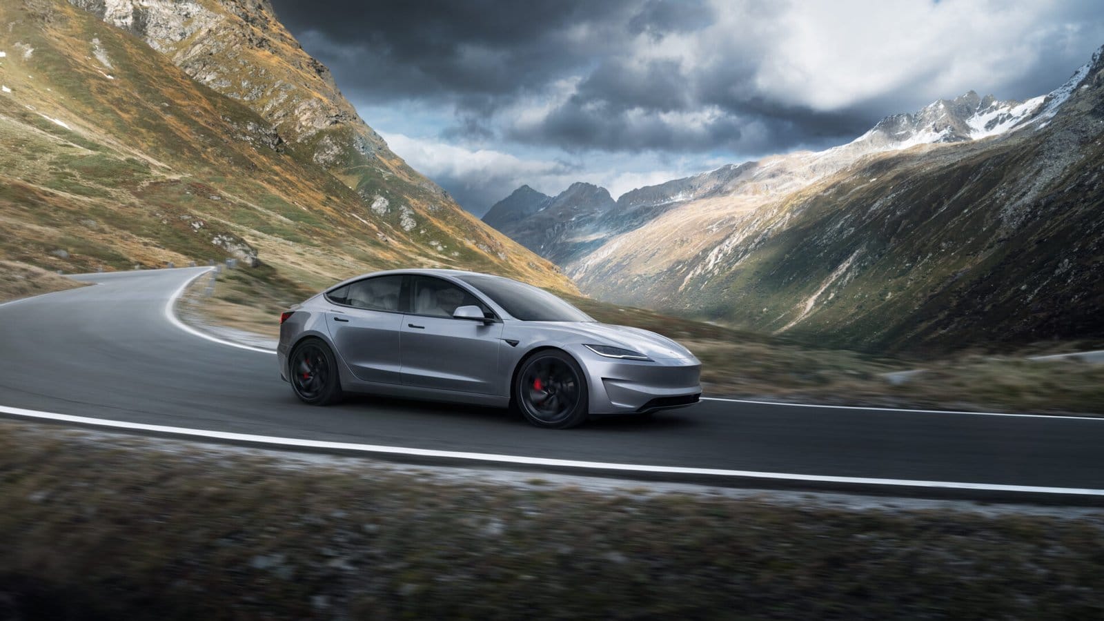 A Tesla Model 3 electric vehicle navigating a winding mountain road under a cloudy sky, surrounded by rugged terrain.