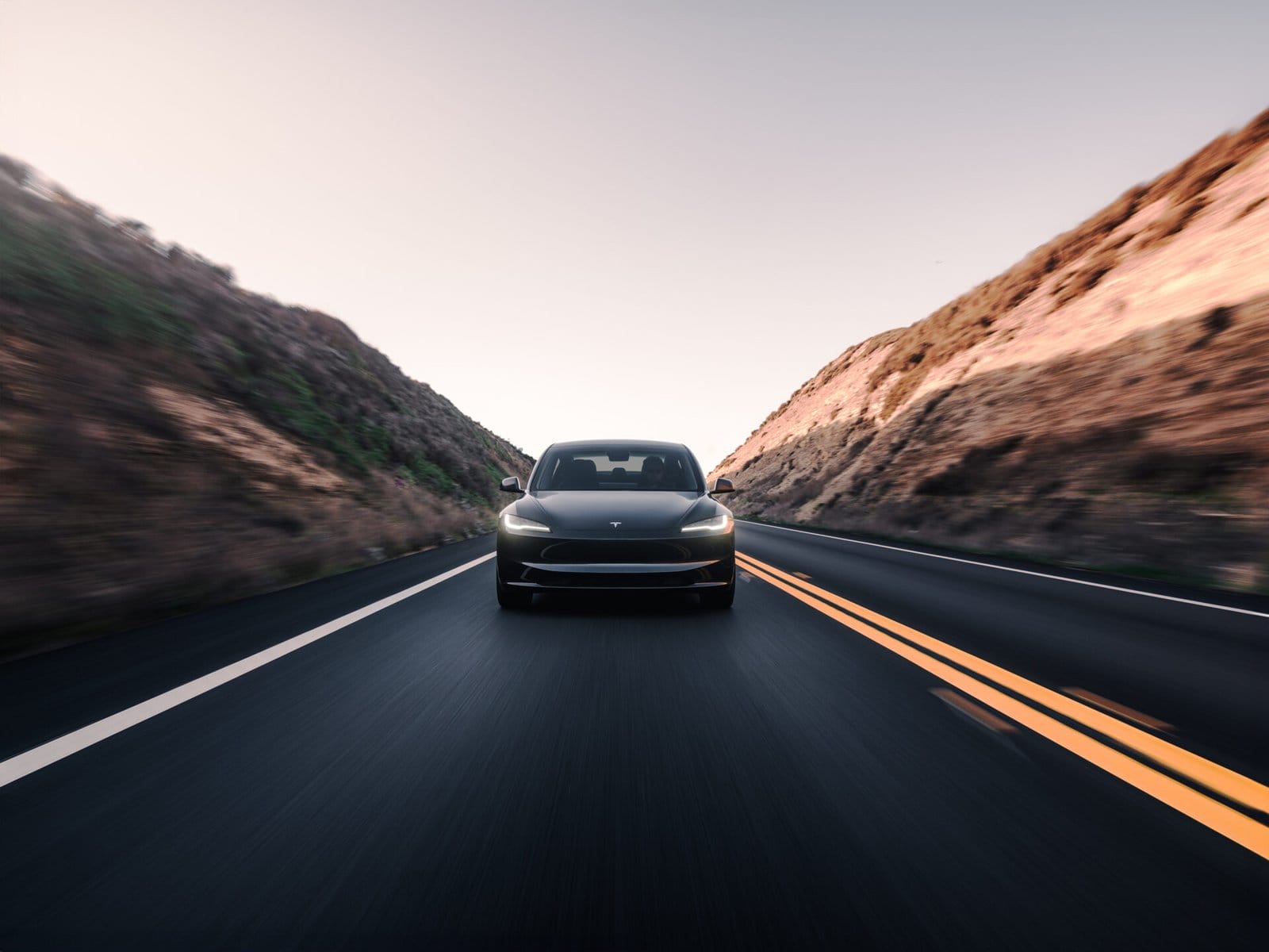 A Tesla Model 3 electric vehicle driving along a scenic highway with hills on both sides, captured from a front-facing perspective.