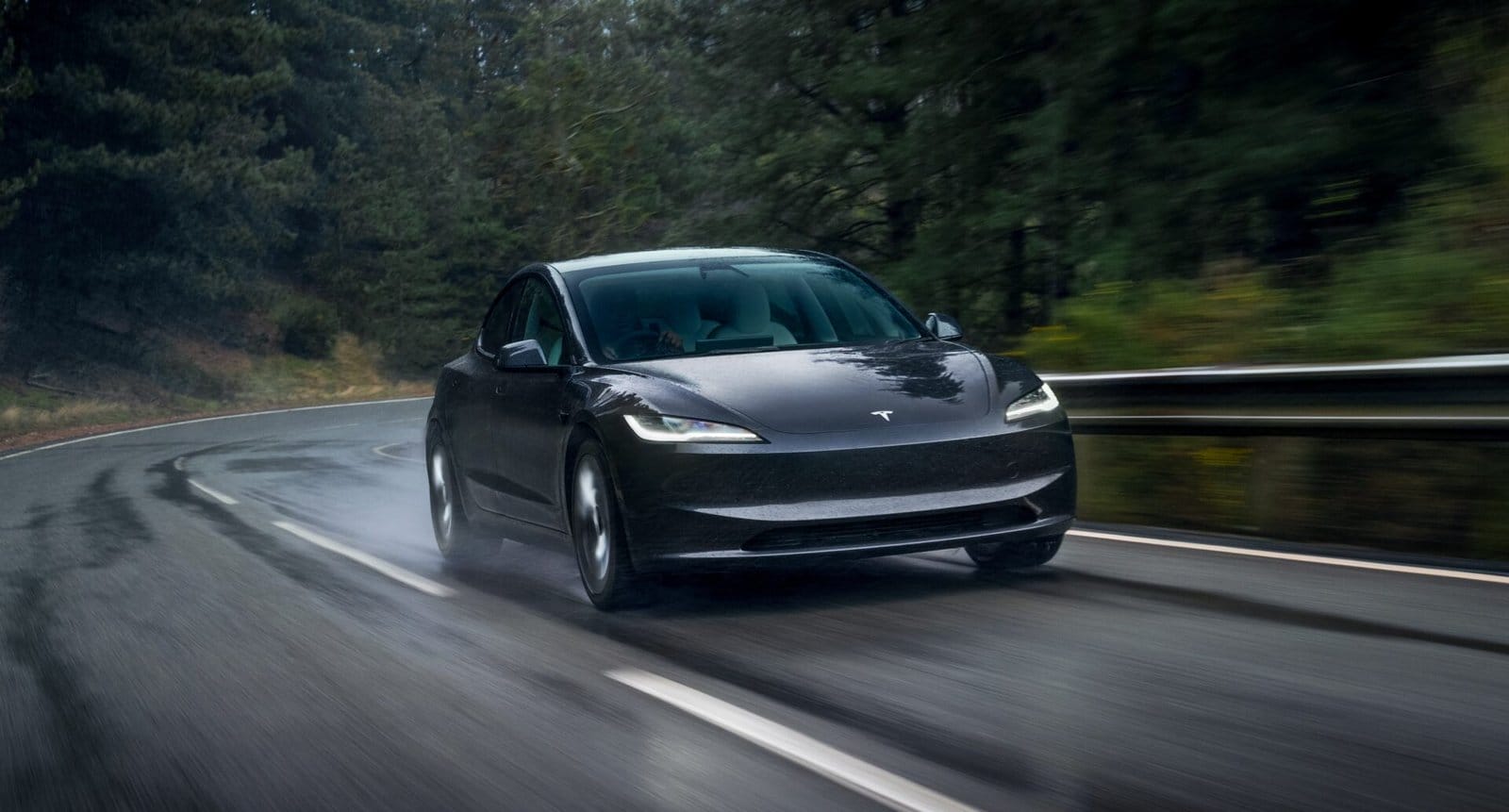 A dark-colored Tesla Model 3 electric vehicle driving on a curvy, wet road surrounded by dense greenery.