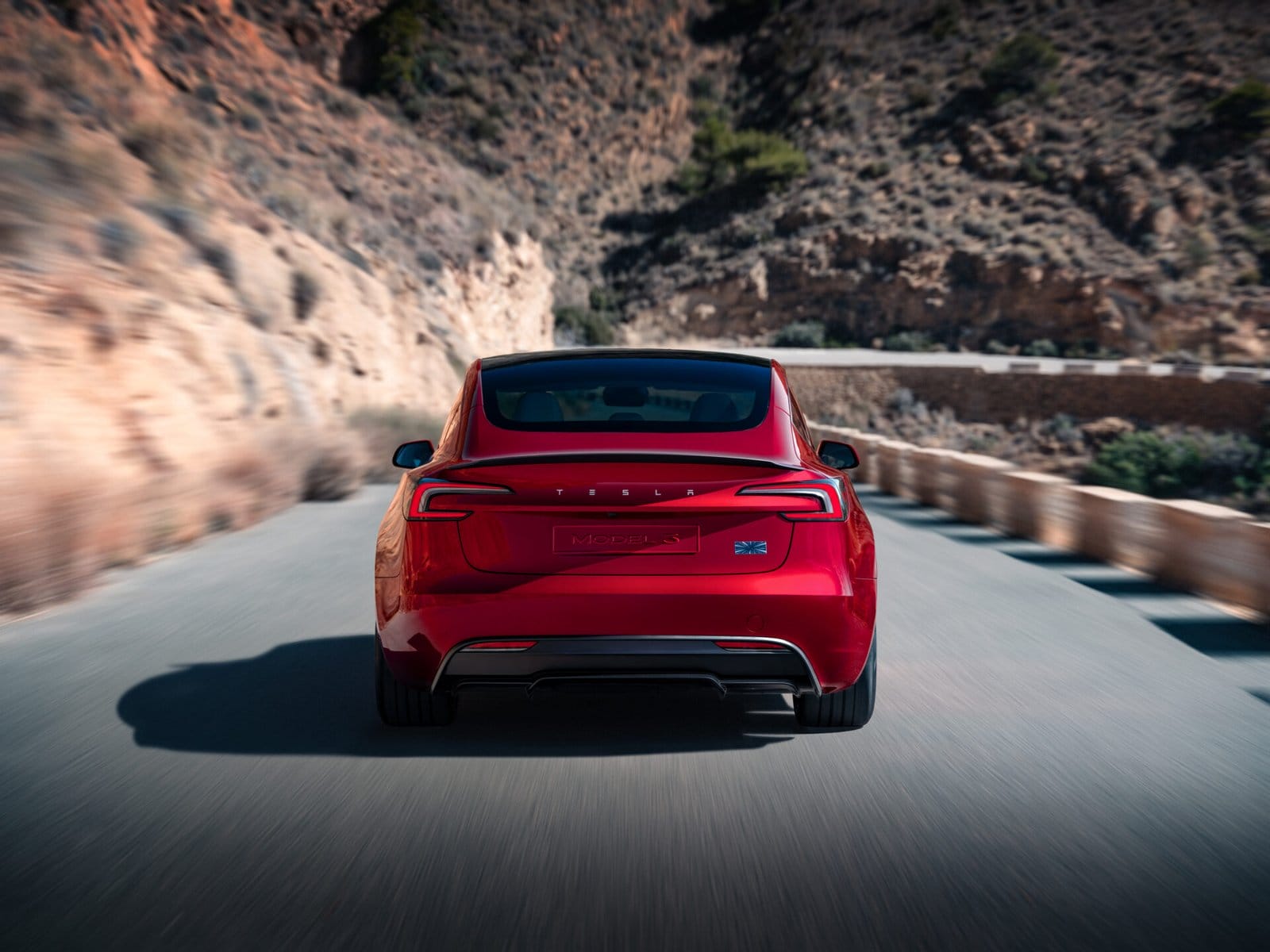 A red Tesla Model 3 electric vehicle captured from the rear as it drives along a winding coastal road with rocky cliffs in the background.