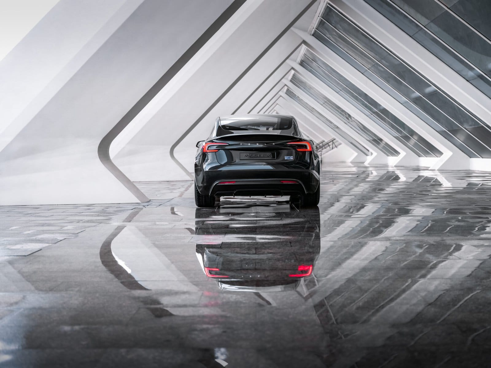 A Tesla Model 3 electric vehicle parked in a futuristic white building, captured from the rear with its taillights reflecting on the polished floor.