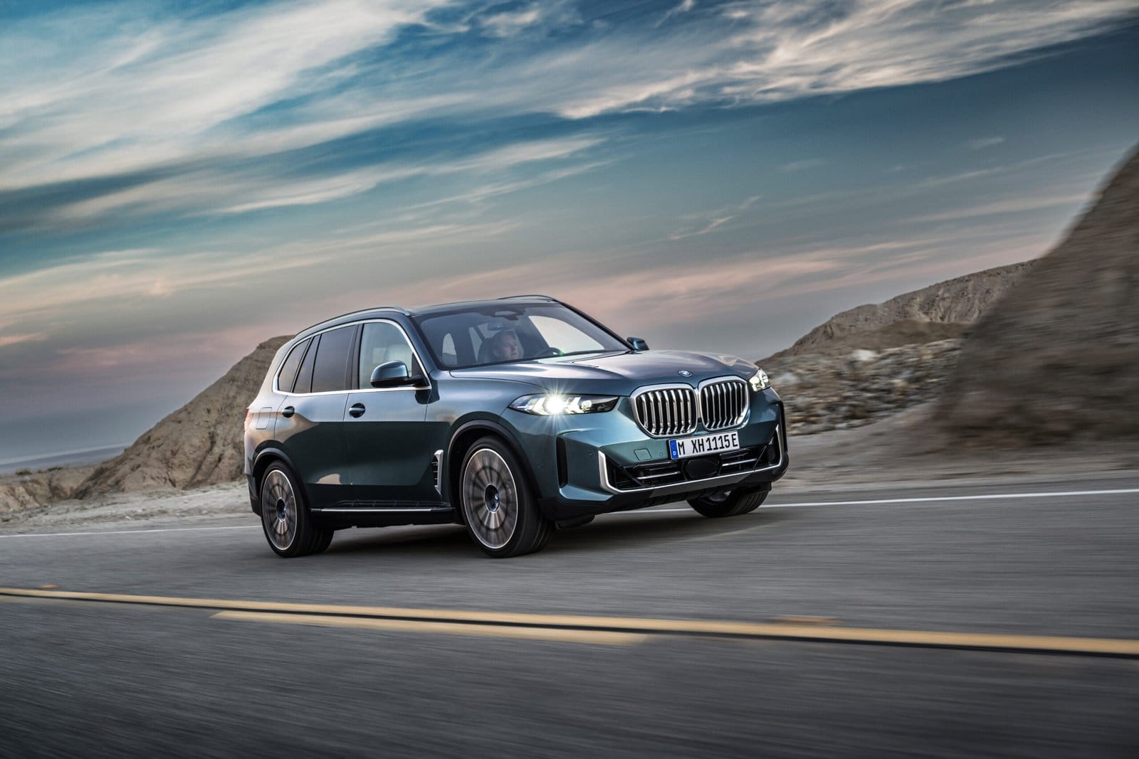 BMW X5 Plug-in Hybrid electric vehicle in motion on a desert road during sunset with rugged hills in the background.