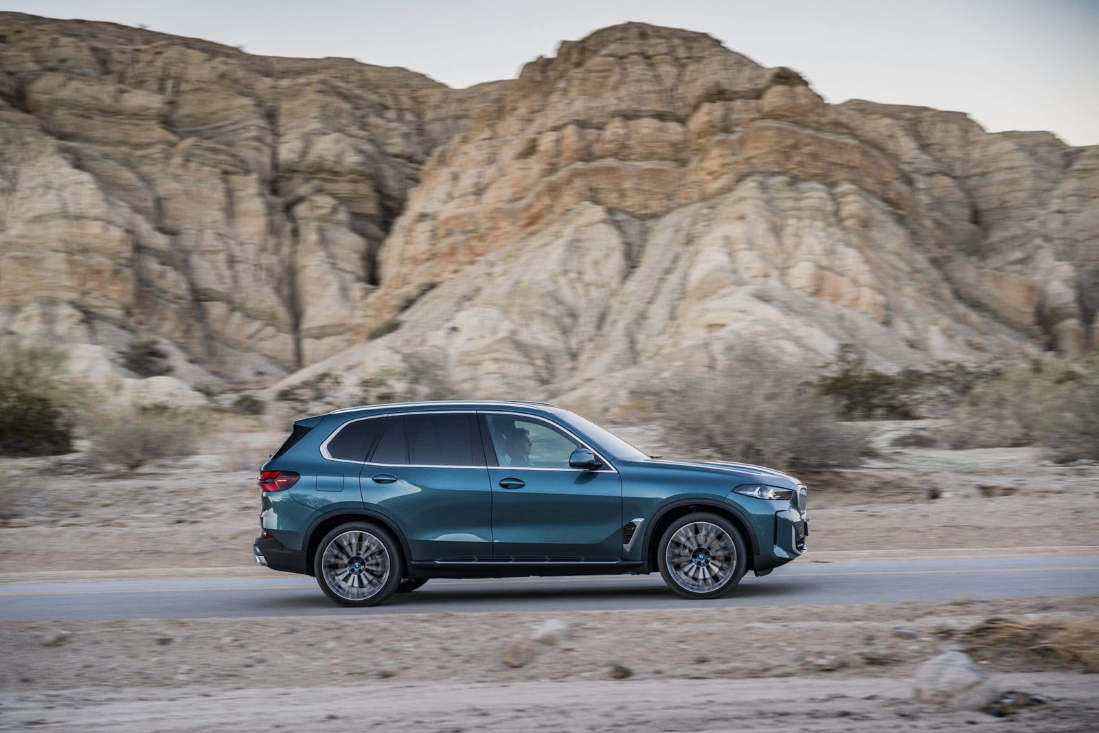 A BMW X5 Plug-in Hybrid electric vehicle driving in profile view along a paved road with towering rock formations in the background.