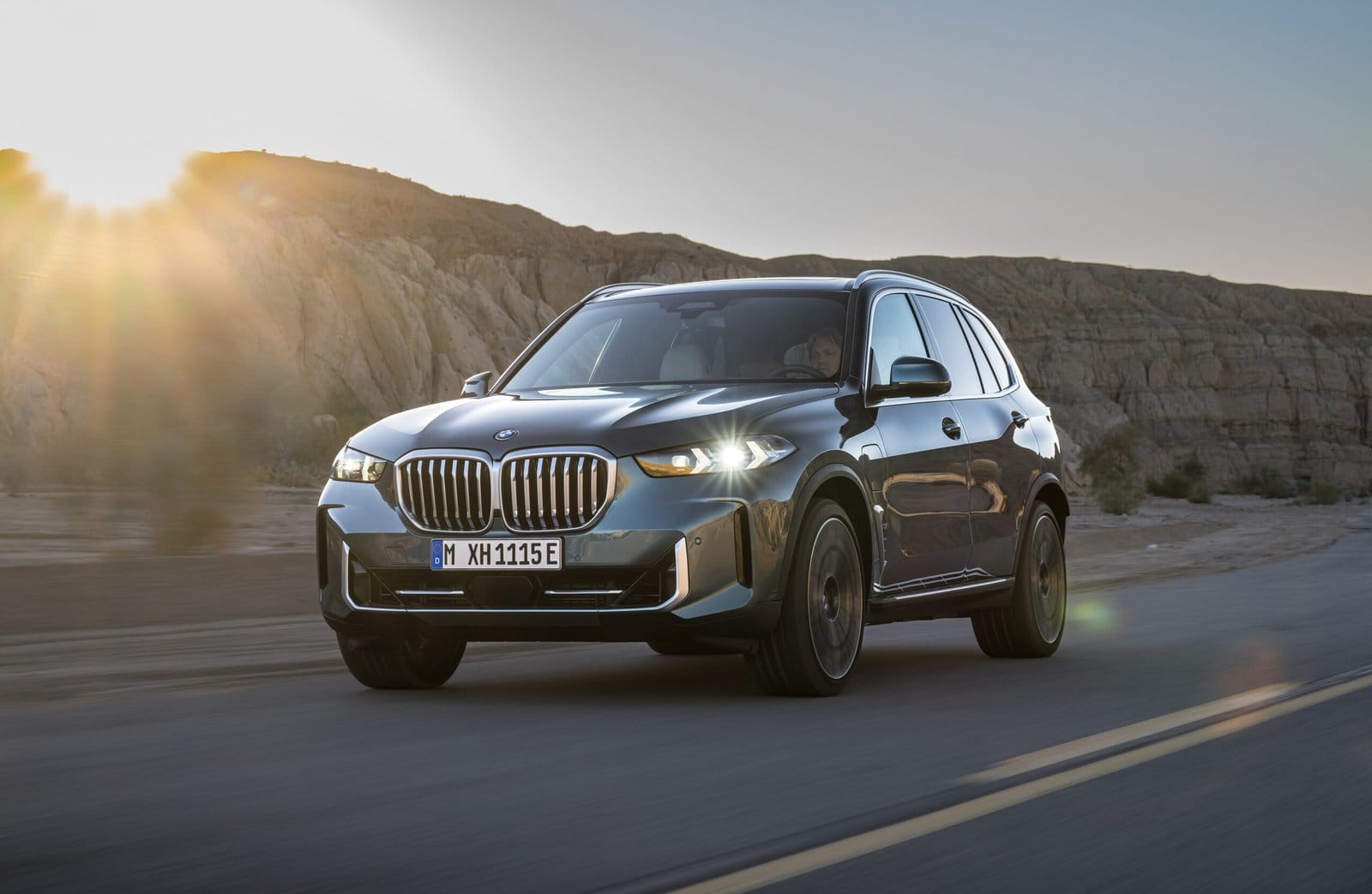 A BMW X5 Plug-in Hybrid electric vehicle driving through a desert landscape with the sun setting behind rocky hills.