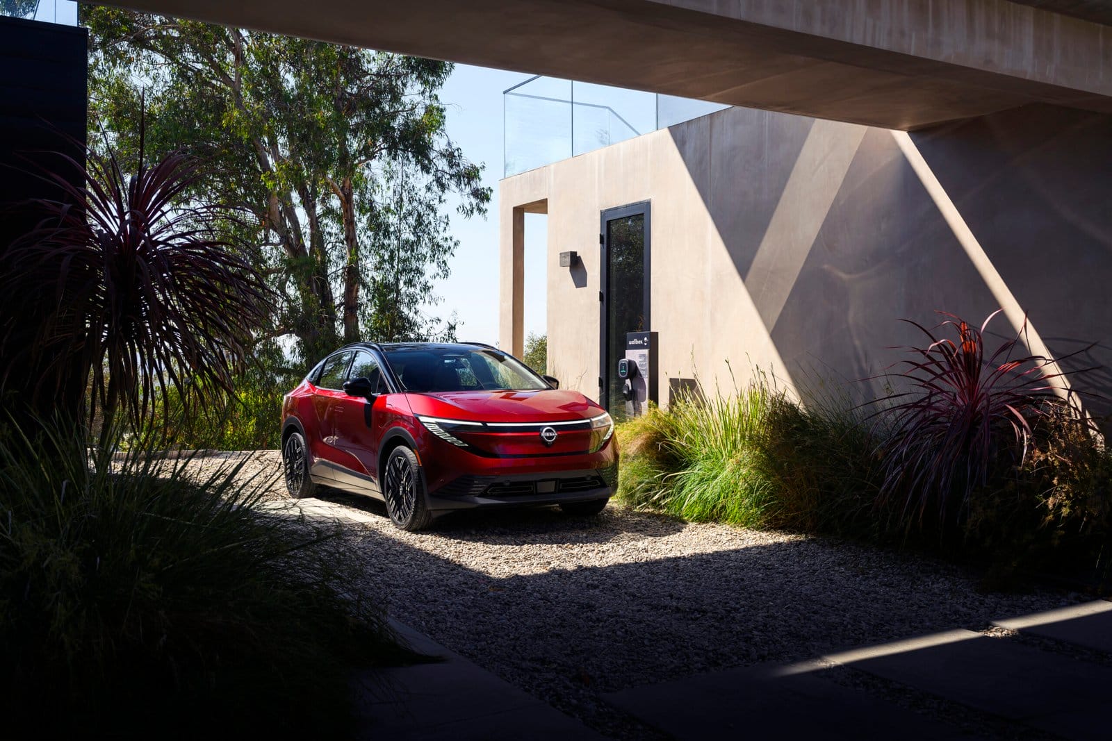 Red Nissan Leaf electric vehicle parked in a modern home's gravel driveway next to an EV charging station