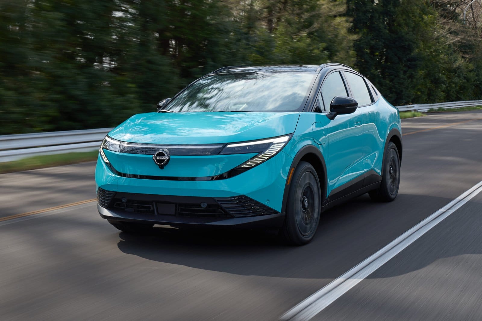Nissan Leaf electric vehicle in blue driving toward the camera on a two-lane road lined with trees