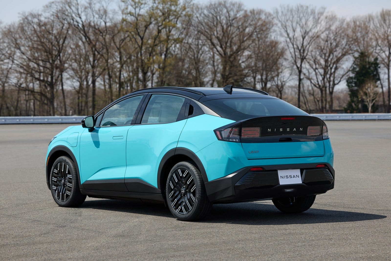 Rear and side view of a parked blue Nissan Leaf electric vehicle in an open paved area with trees in the background