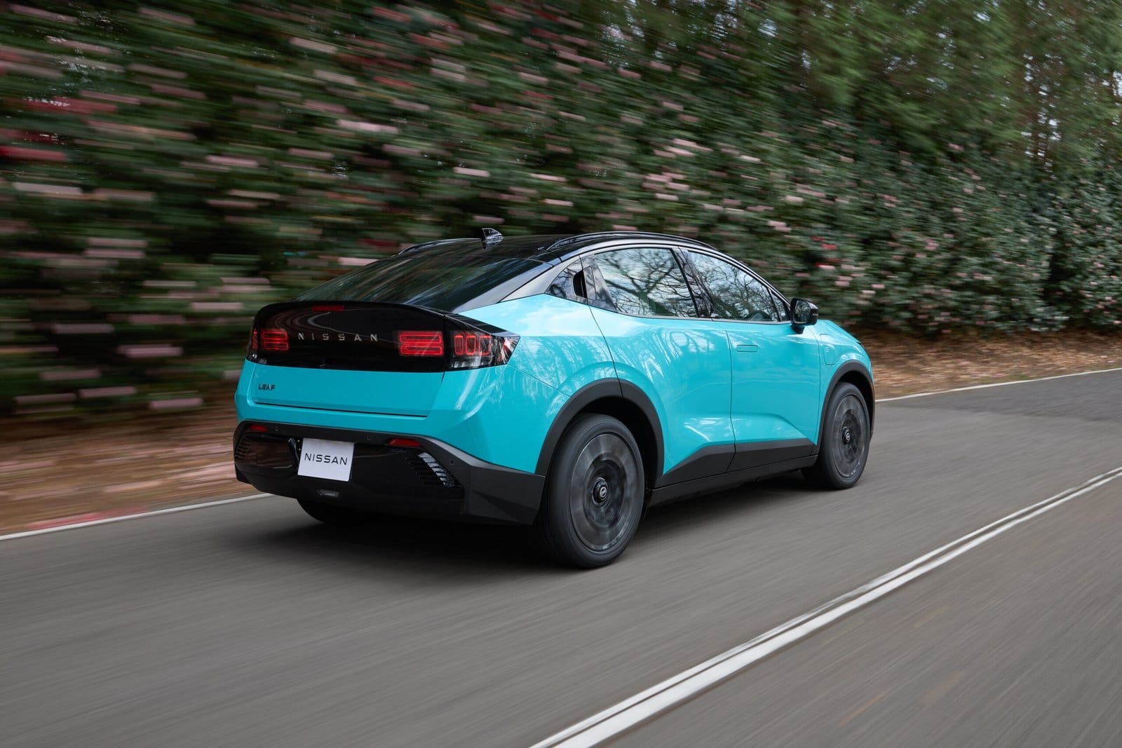 Rear and side view of a parked blue Nissan Leaf electric vehicle in an open paved area with trees in the background