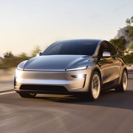 A Tesla Model Y electric vehicle drives along a winding road at sunset, with headlights on and a tunnel in the background.