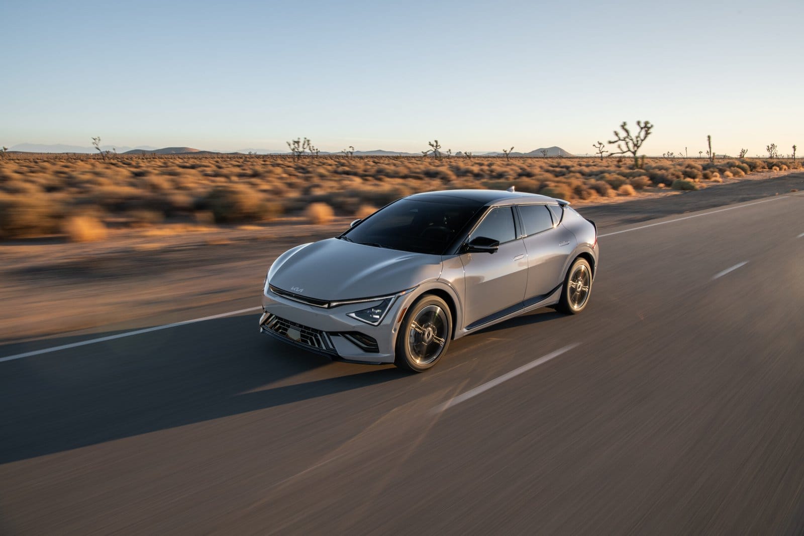A silver Kia EV6 electric vehicle driving along a straight desert road at sunset, with Joshua trees and mountains in the background.