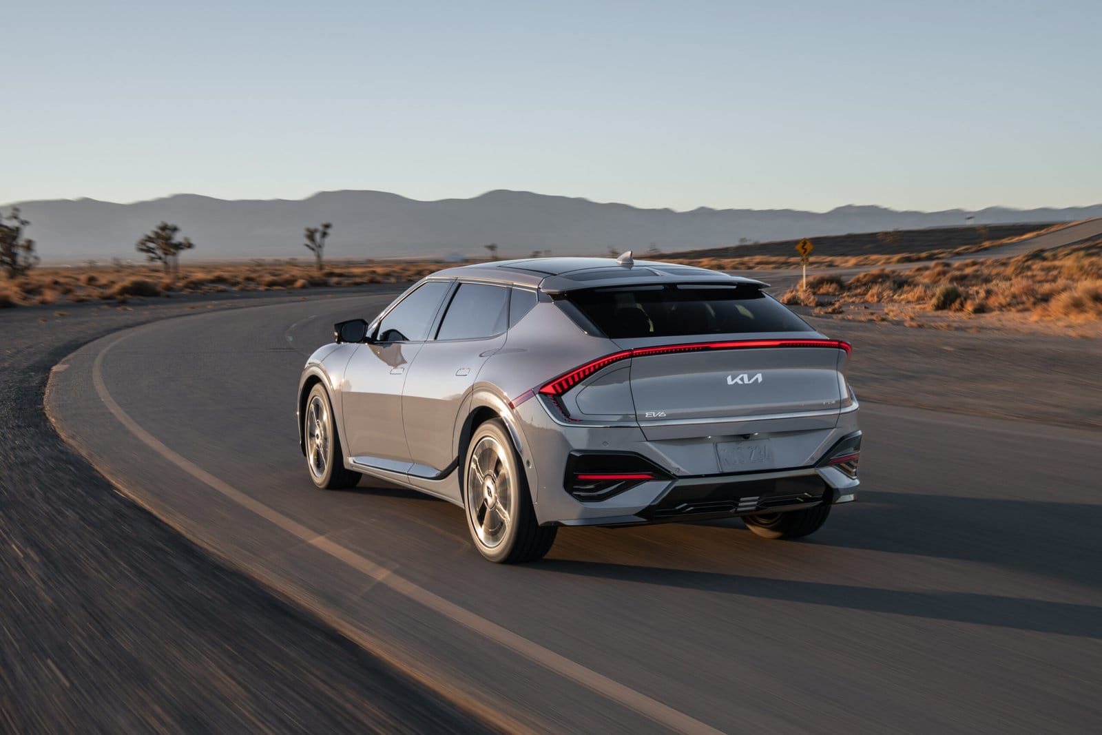 A silver Kia EV6 electric vehicle moving along a winding desert road with mountains in the background at sunset.