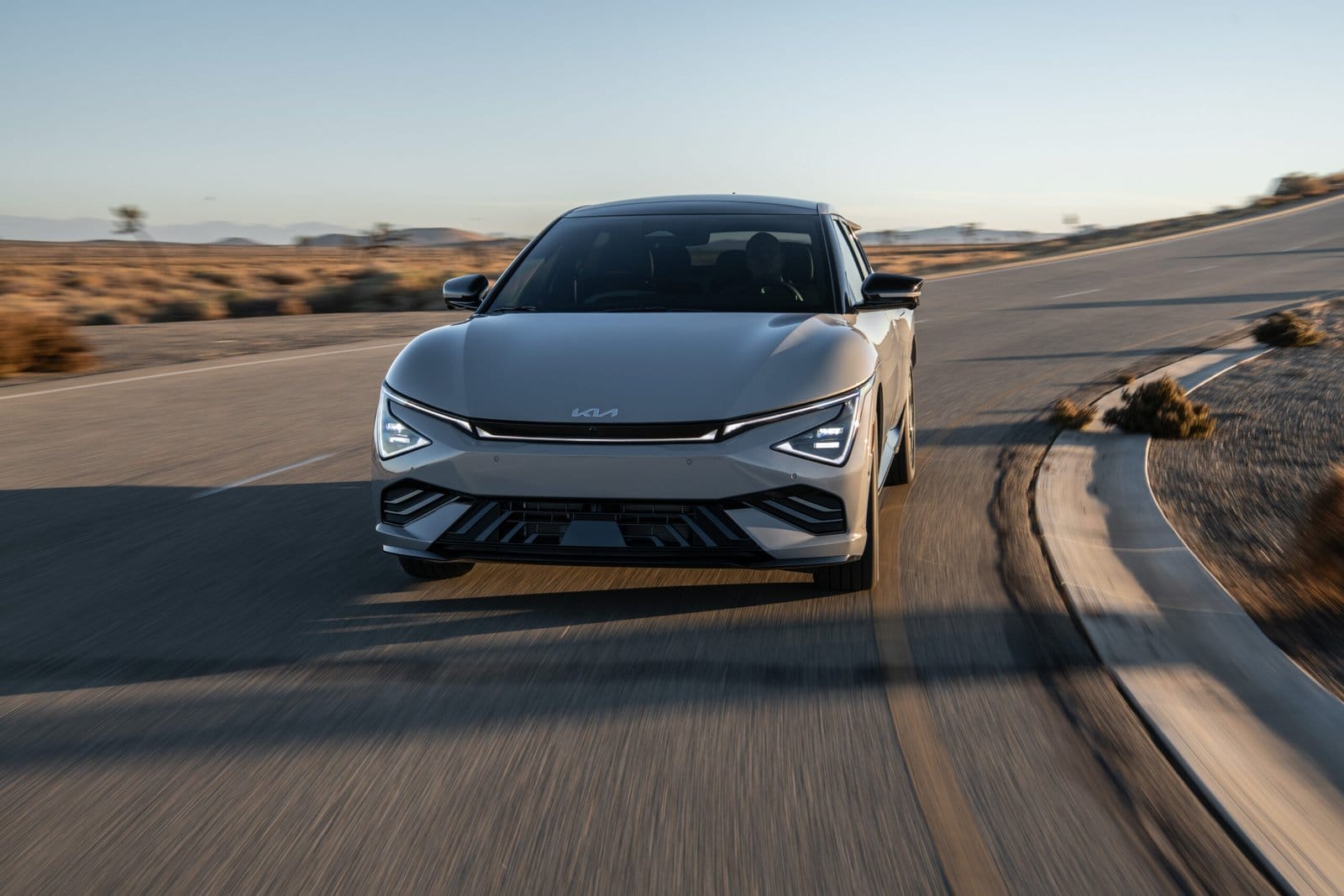 A silver Kia EV6 electric vehicle captured from the front, driving on a curved desert highway at sunset.