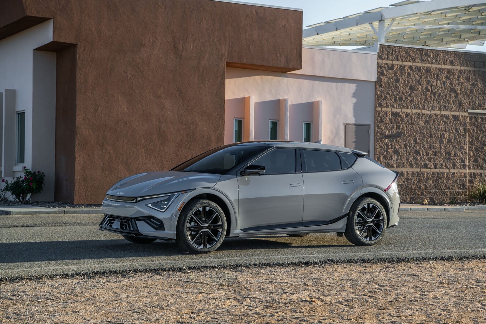 A silver Kia EV6 electric vehicle parked on a roadside near a modern building with earthy tones and solar panel structures.