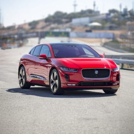 A red Jaguar I Pace electric vehicle driving on a track with a blurred industrial and hilly background.