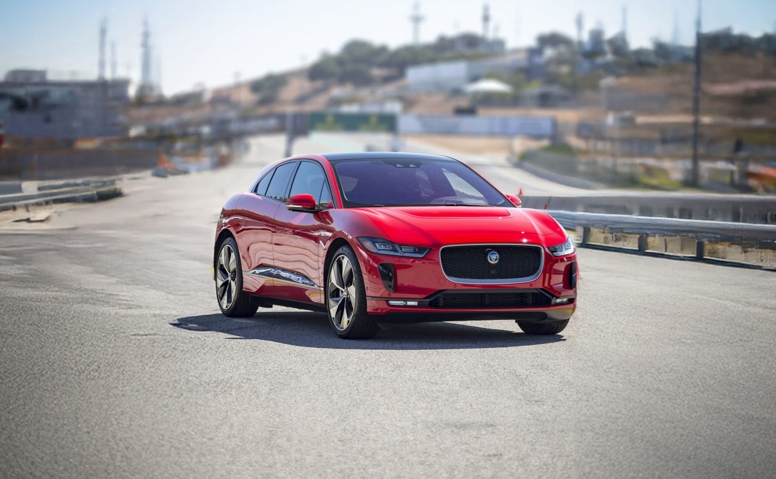 A red Jaguar I Pace electric vehicle driving on a track with a blurred industrial and hilly background.