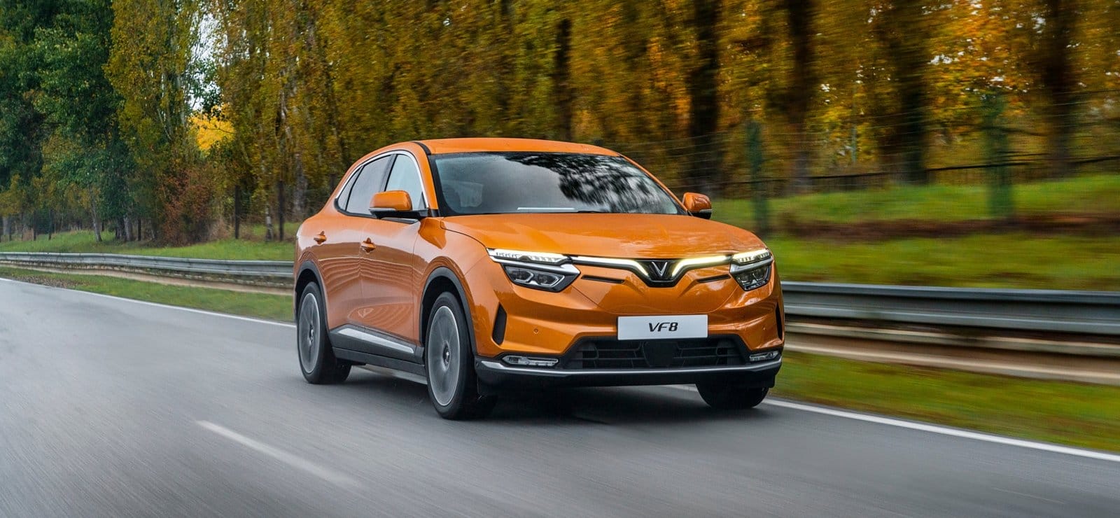 Bright orange Vinfast VF8 electric vehicle driving on a forest road surrounded by autumn trees