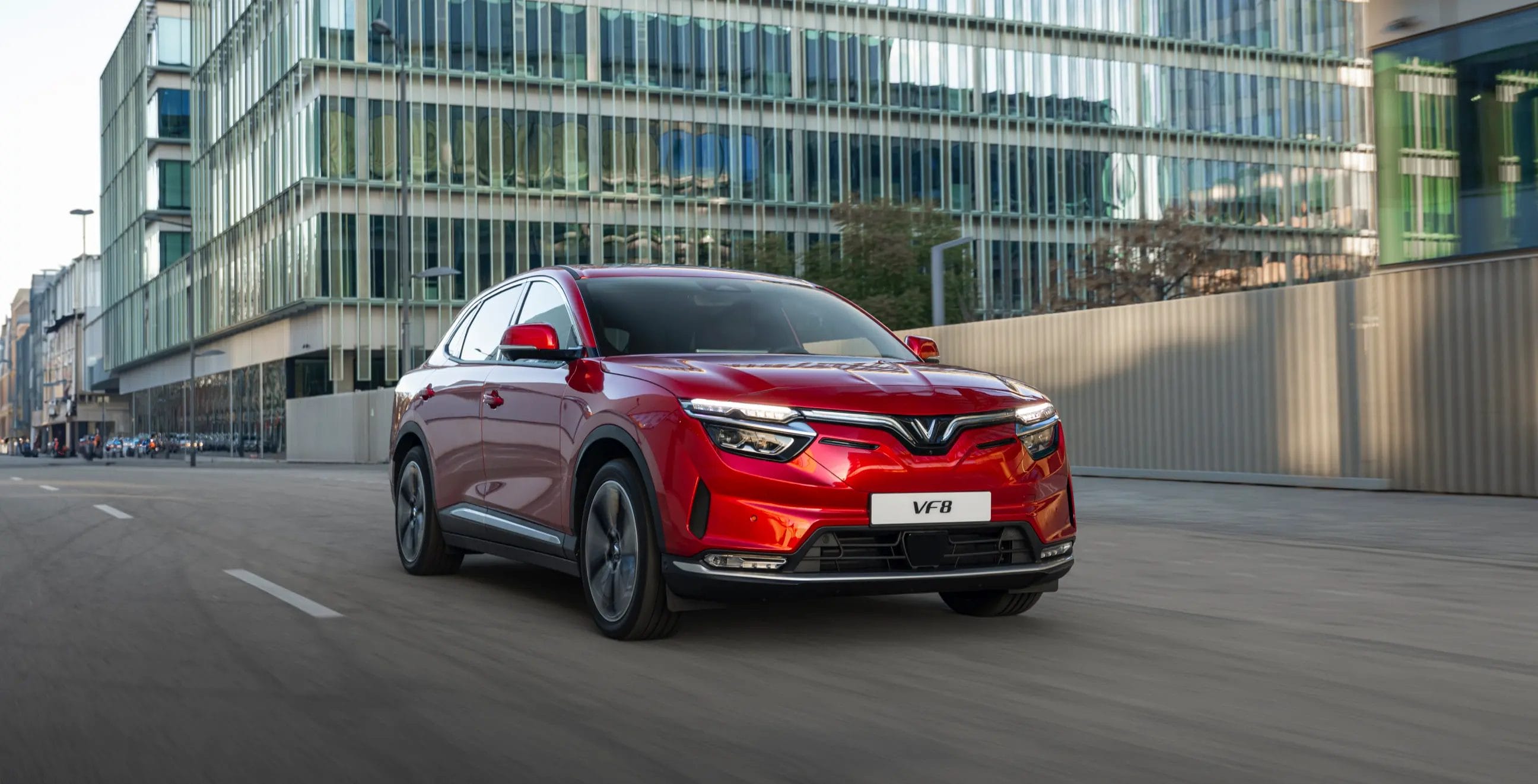 Front side view of a red Vinfast VF8 electric vehicle driving past tall modern office buildings in the city