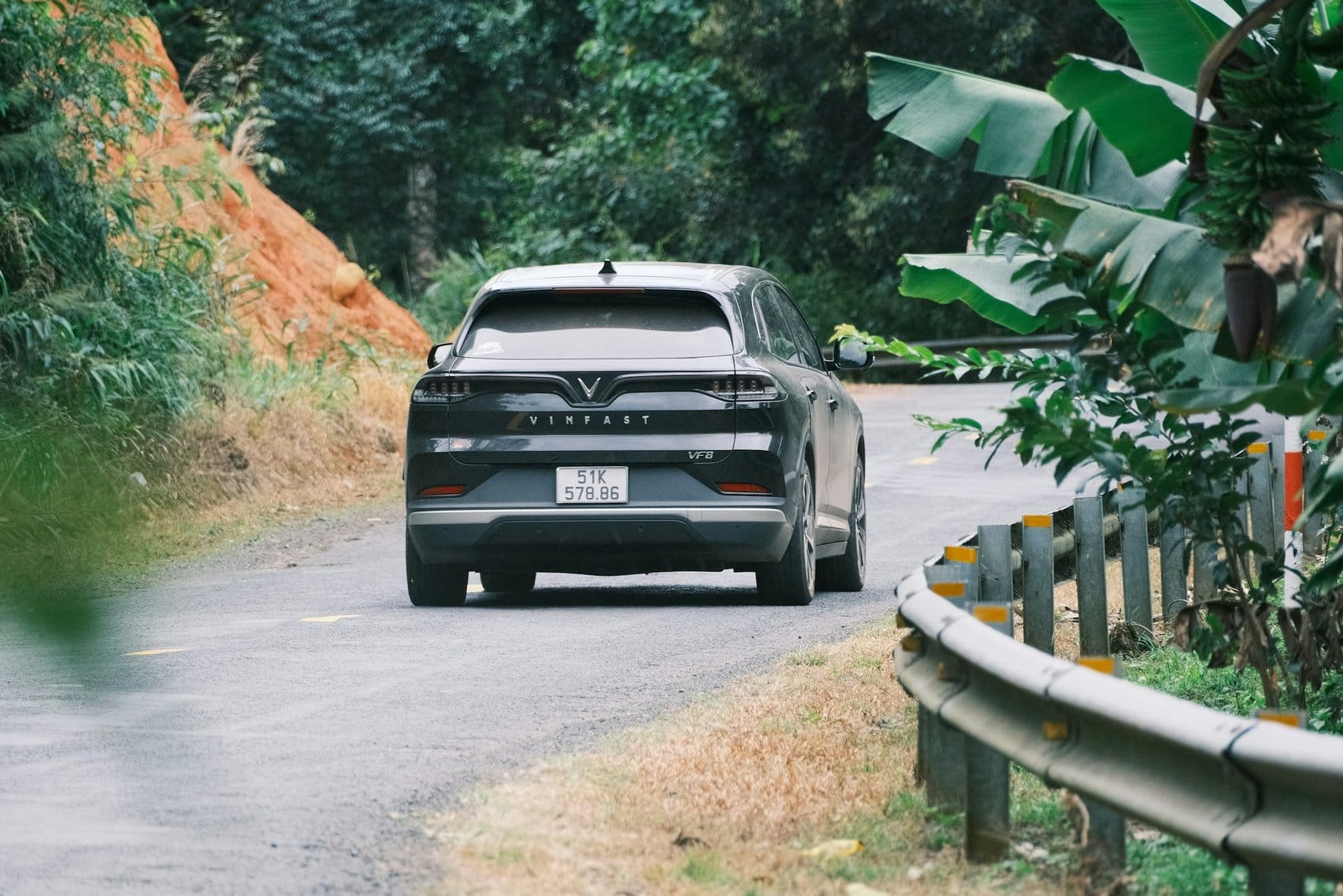 Rear view of a black Vinfast VF8 electric vehicle on a curved forest road surrounded by greenery