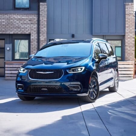 A blue Chrysler Pacifica electric vehicle parked in a modern residential driveway with brick and glass elements in the background.