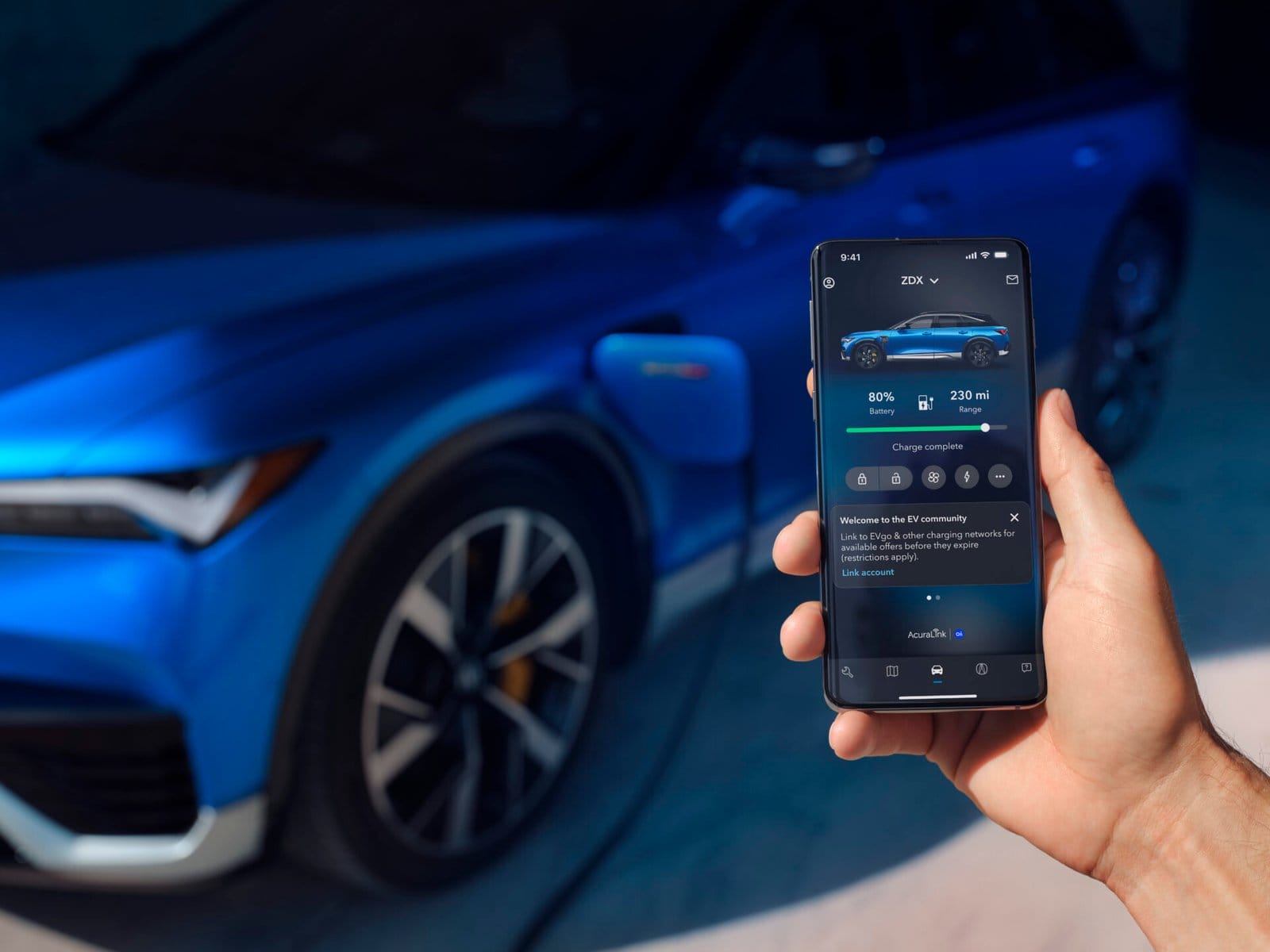 A hand holding a smartphone displaying the charging status of a blue Acura ZDX electric vehicle, which is connected to a charger in the background.