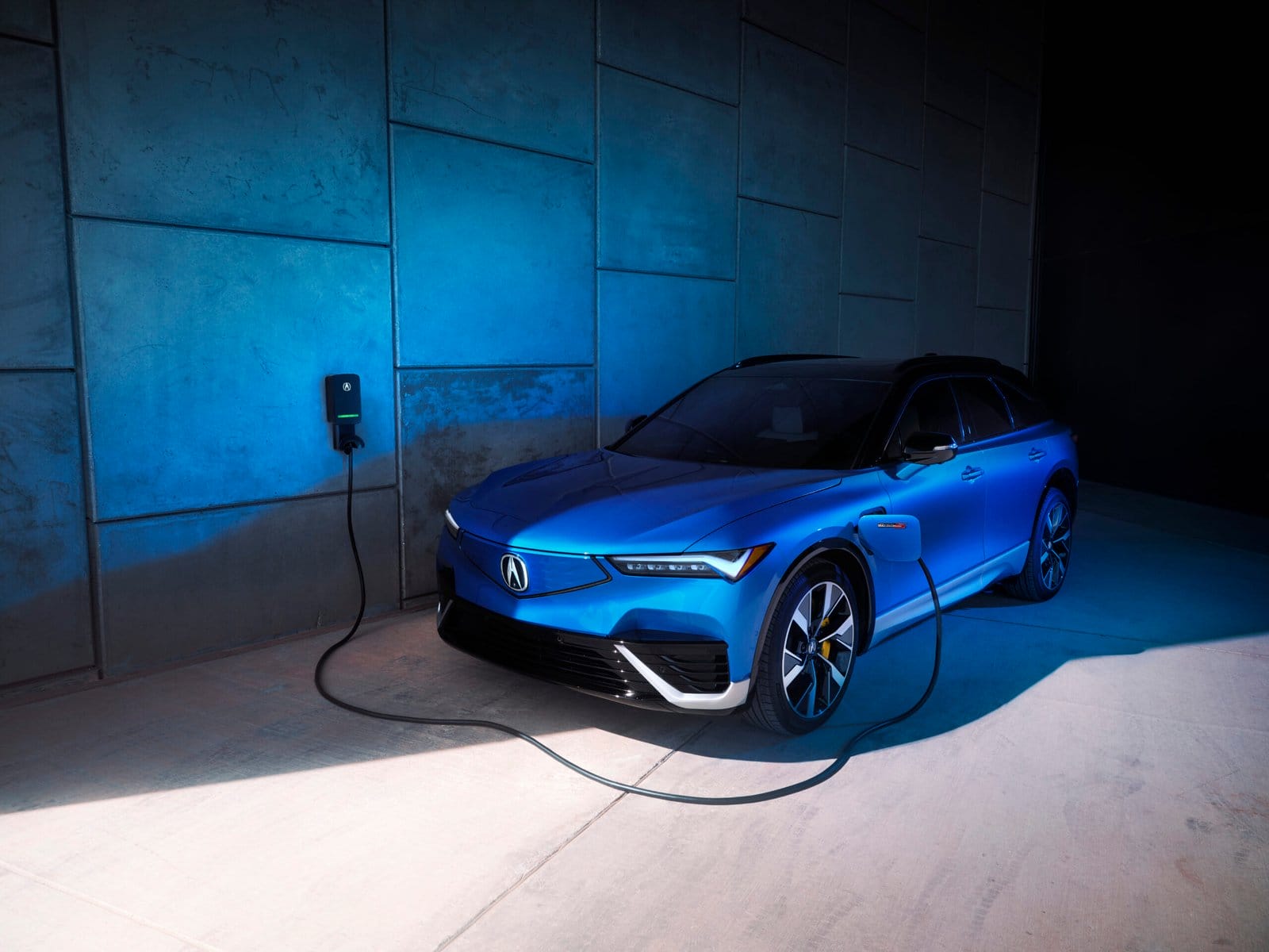 A blue Acura ZDX electric vehicle plugged into a wall-mounted charging station in a dimly lit space with blue lighting.