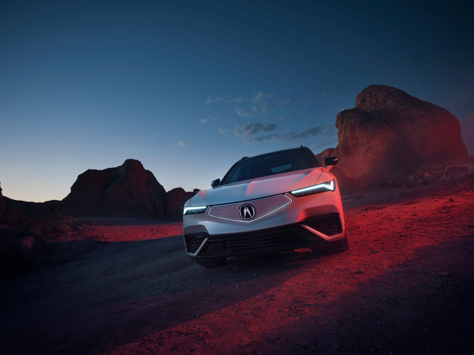 Front view of a silver Acura ZDX electric vehicle parked on a rugged desert terrain at dusk, surrounded by rocky formations under a twilight sky.