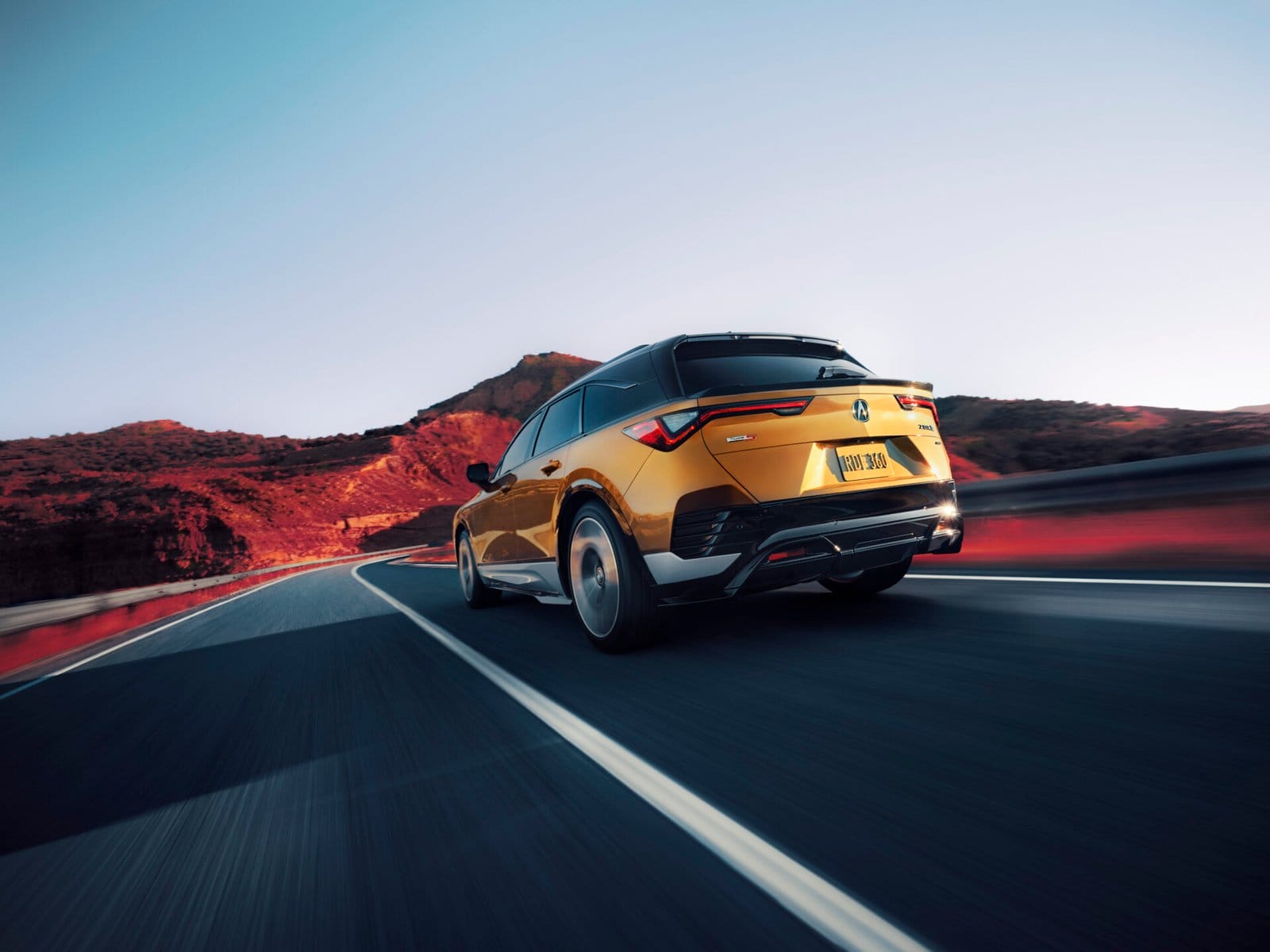 Rear view of a golden Acura ZDX electric vehicle driving along a winding road with red hills in the background during sunset.