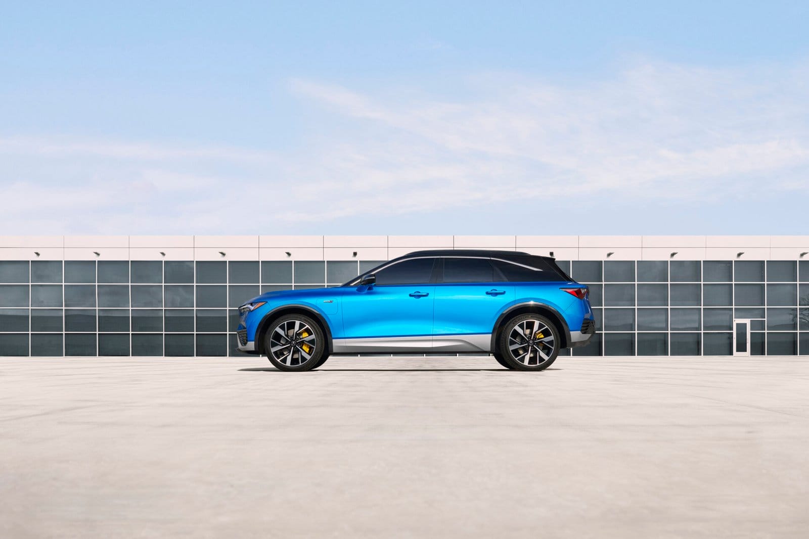 Side view of a blue Acura ZDX electric vehicle parked in front of a modern building with a clear sky background.
