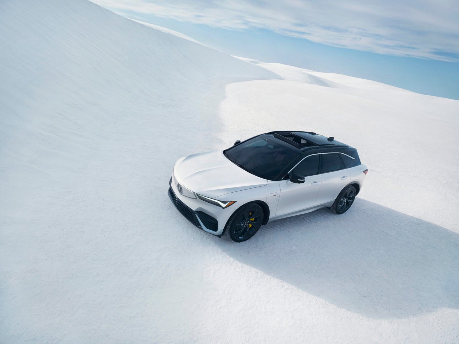 A white Acura ZDX electric vehicle parked on an expansive snow-covered terrain under a bright blue sky, showcasing its sleek design and versatility.
