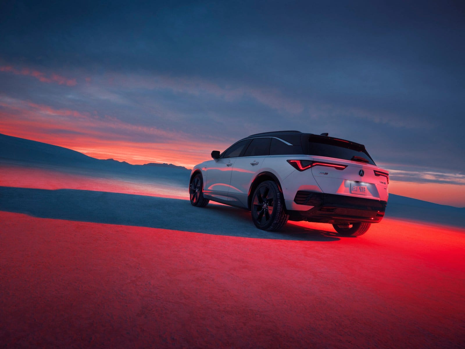 Rear view of a white Acura ZDX electric vehicle parked on a flat terrain during a vibrant sunset, with mountains in the distance and a glowing sky.