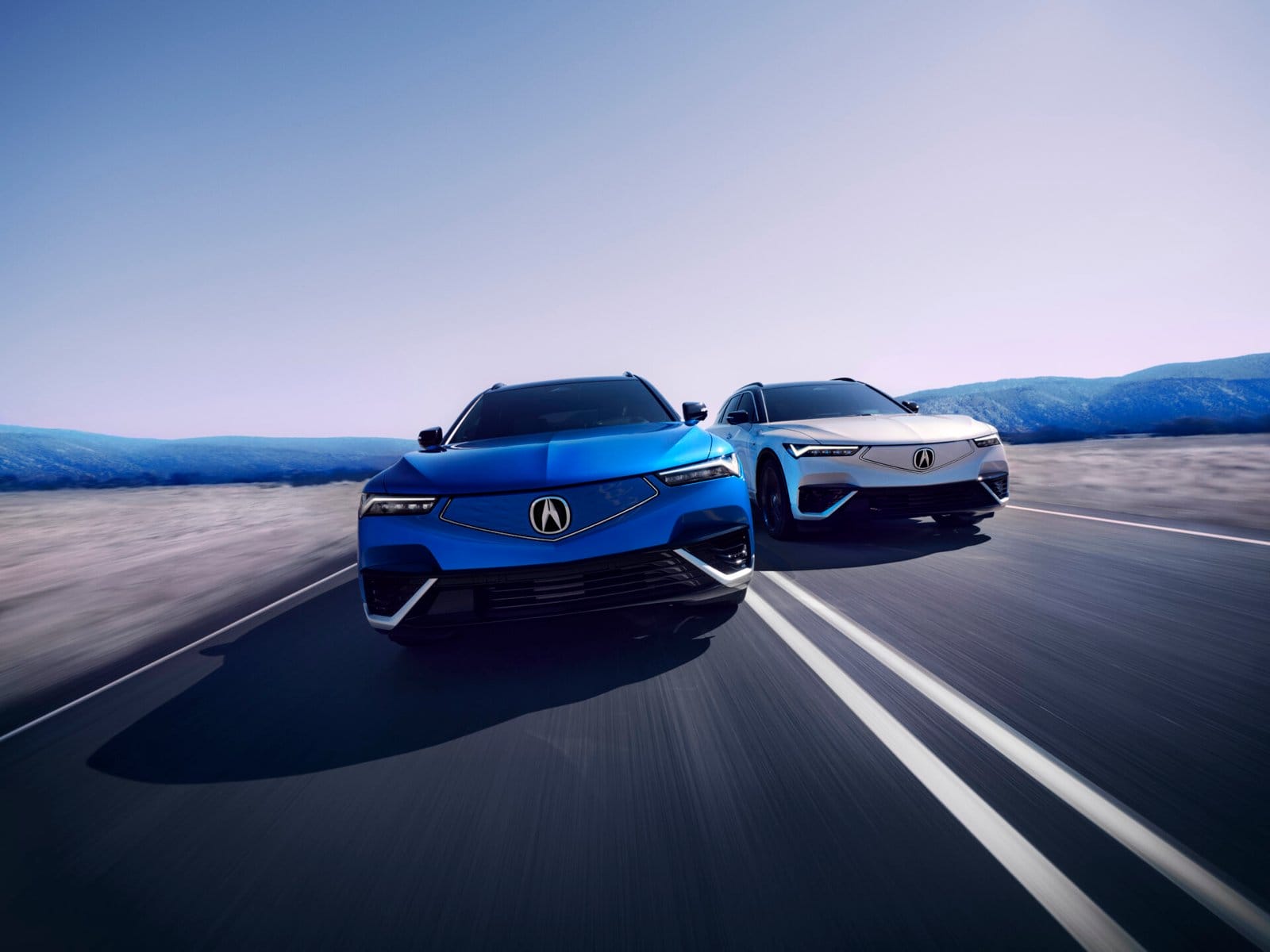 Front view of two Acura ZDX electric vehicles, one blue and one silver, driving side by side on a wide road with a scenic mountain background.