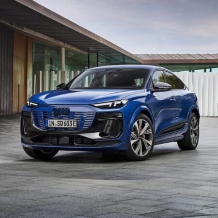 A blue Audi SQ6 e-tron electric vehicle parked in a modern courtyard with clean tiled flooring, a concrete wall, and a white minimalist building in the background.