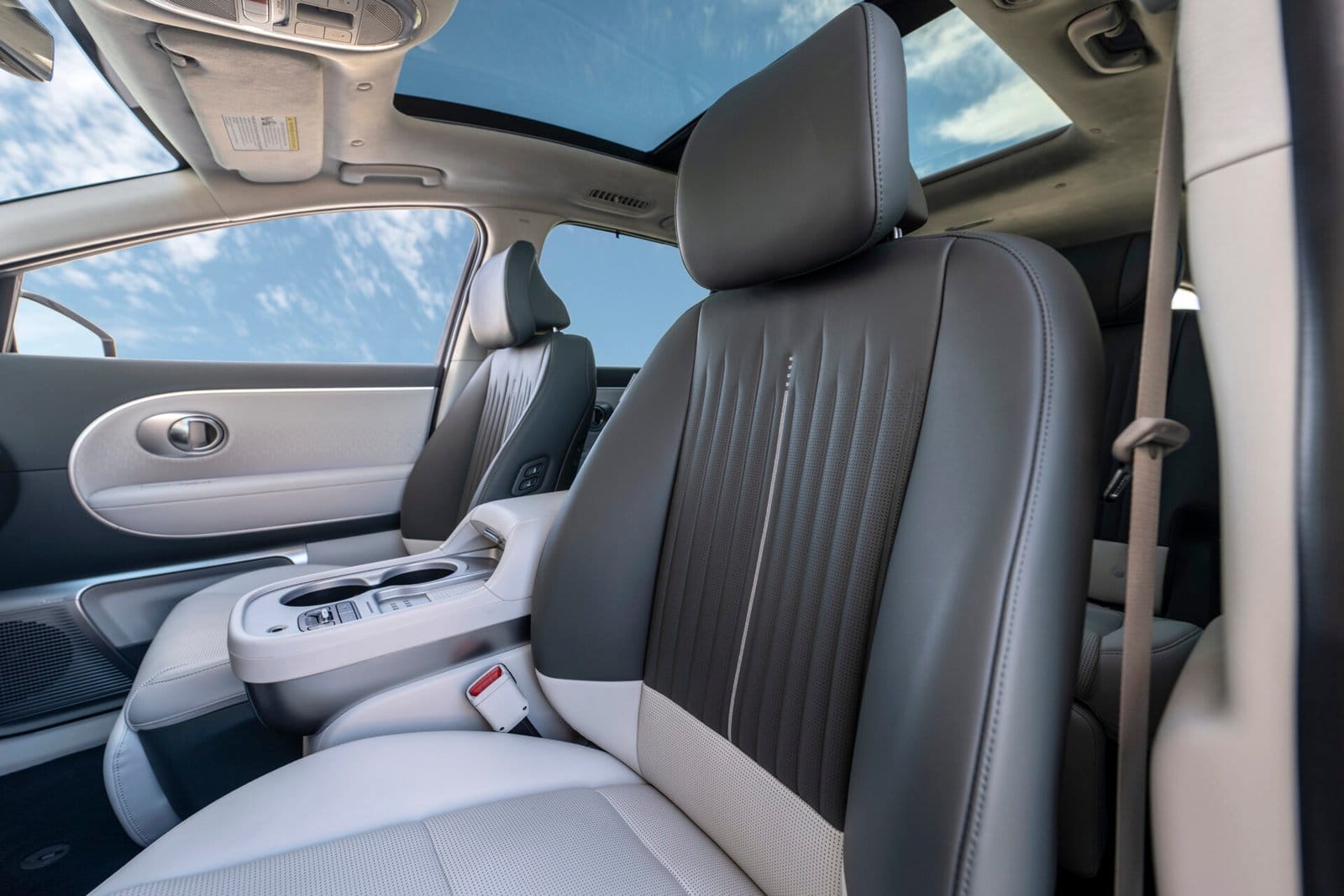 A view of the Hyundai Ioniq 9 electric vehicle's front seats with gray and white upholstery, complemented by a panoramic sunroof and a bright blue sky visible through the glass.