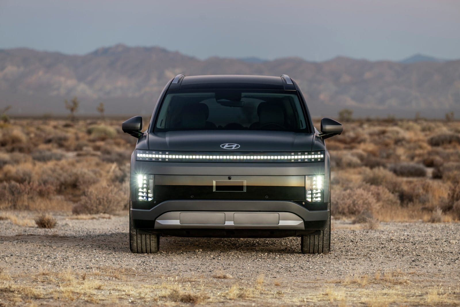 The Hyundai Ioniq 9 electric vehicle seen from the front, showcasing its illuminated pixel-style headlights and grille design, set in a dry desert landscape with distant mountains.