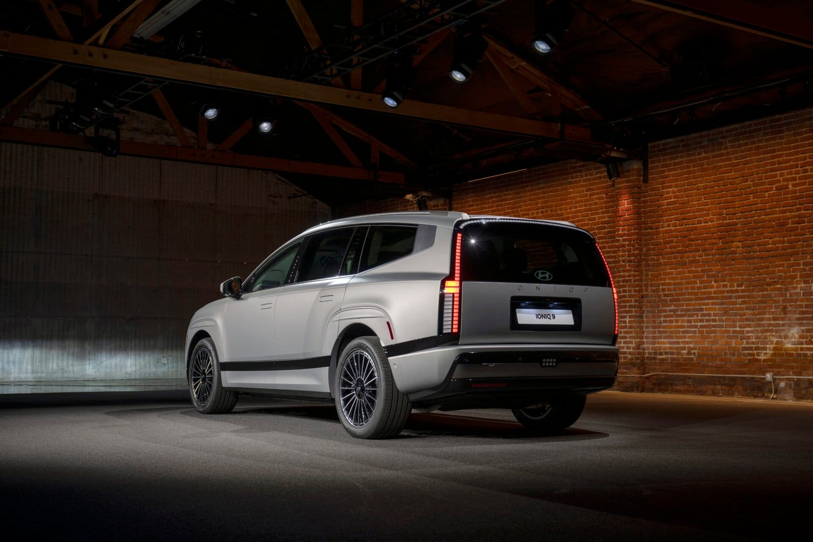 Rear three-quarter view of the Hyundai Ioniq 9 electric vehicle under showroom lighting, highlighting its sleek design, vertical tail lights, and alloy wheels. The brick and industrial backdrop adds contrast to the vehicle's modern styling.