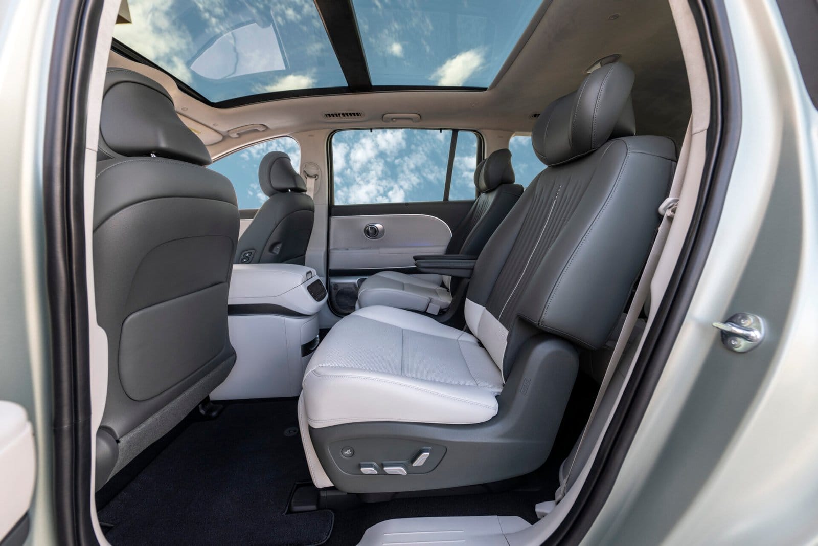 The rear cabin of the Hyundai Ioniq 9 electric vehicle, featuring two-tone reclining seats with adjustable controls and a panoramic sunroof that frames a bright blue sky with scattered clouds.