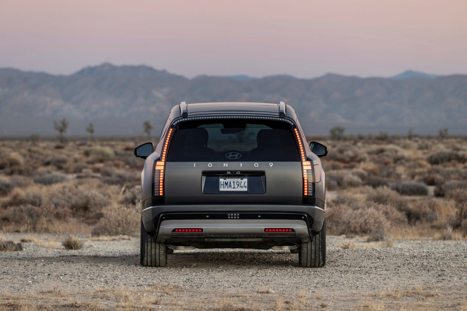The Hyundai Ioniq 9 electric vehicle shown from the rear, highlighting its vertical tail lights and clean design. The vehicle is parked in a desert setting with mountains visible under a soft pink sky at dusk.