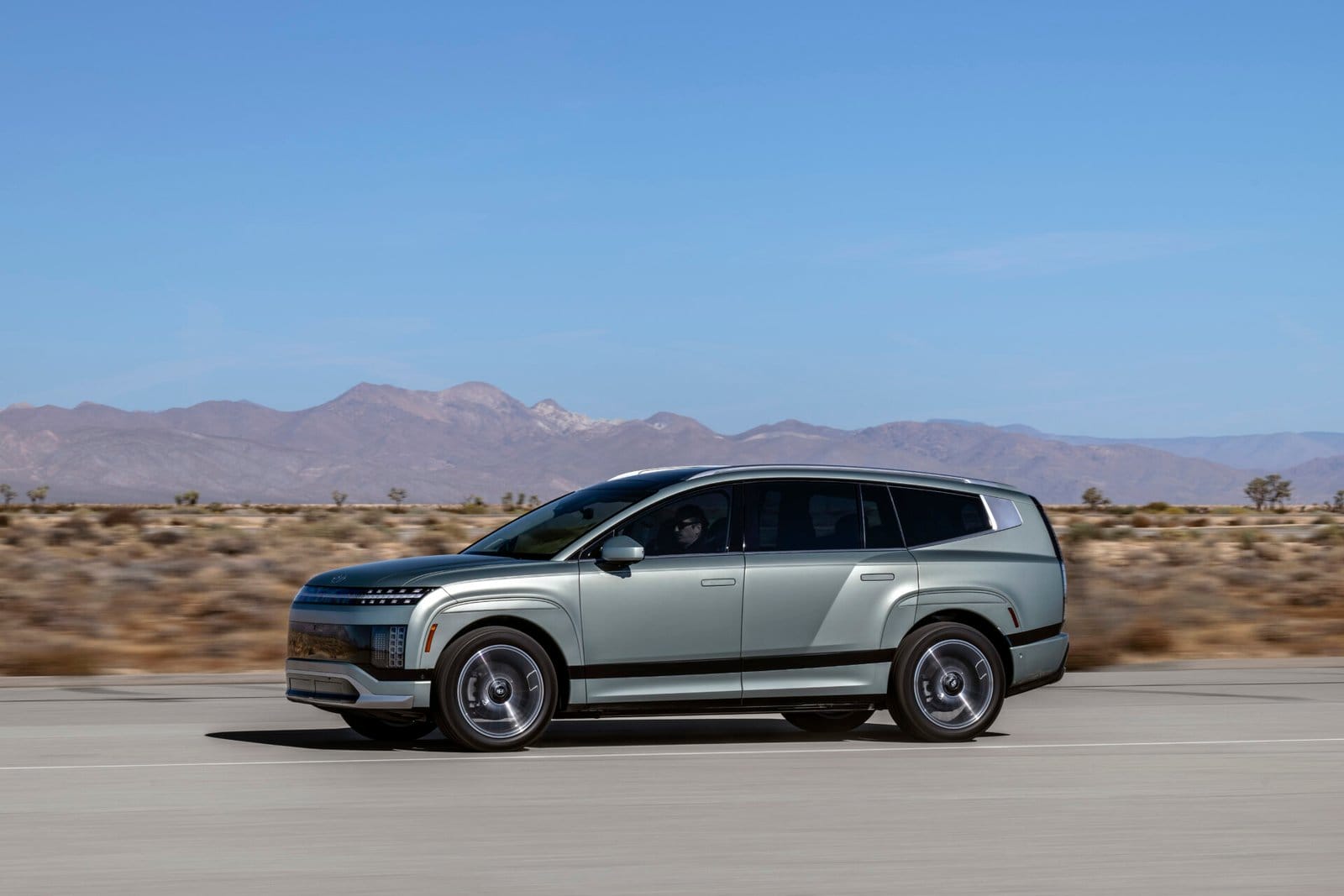 The Hyundai Ioniq 9 electric vehicle captured in motion from a side profile on a paved road, surrounded by desert vegetation and distant mountains under a clear blue sky.