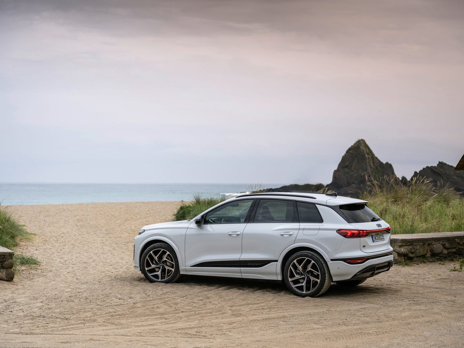 Side view of Audi Q6 electric vehicle parked on sand near beach and rocks