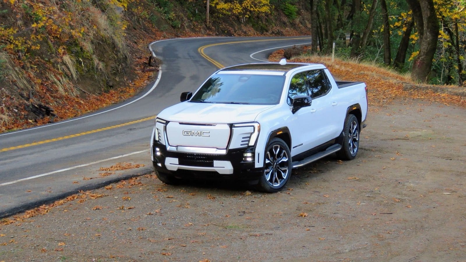 GMC Sierra EV electric vehicle parked near a winding road in a forested area during autumn