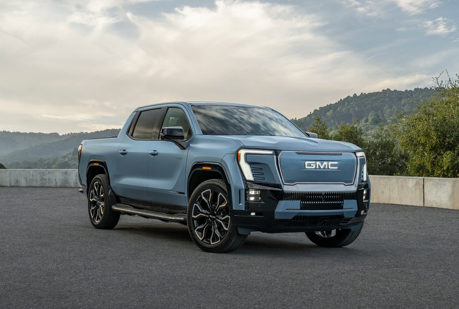 Front three-quarter view of the GMC Sierra EV Denali electric vehicle in blue parked on asphalt with forested hills in the background