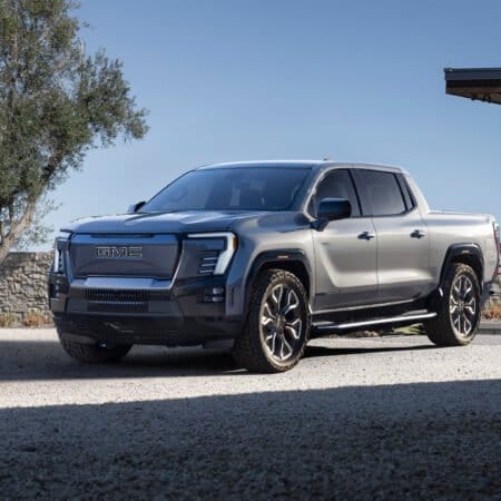Side-front angle of the GMC Sierra EV Denali electric vehicle parked on a gravel driveway near a modern house