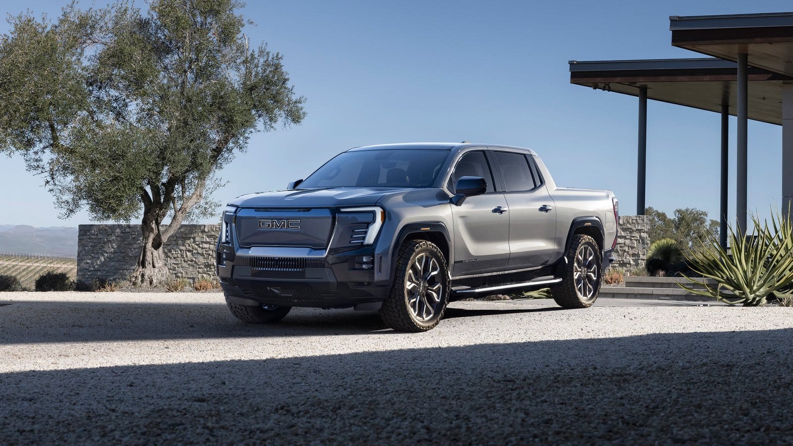 Side-front angle of the GMC Sierra EV Denali electric vehicle parked on a gravel driveway near a modern house