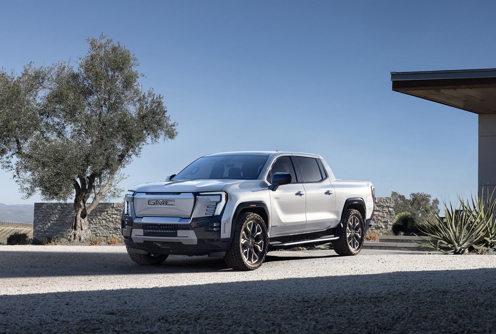 Silver GMC Sierra EV Denali electric vehicle parked outside a contemporary house with desert landscaping and blue sky background