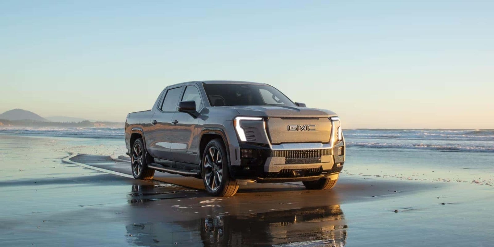 GMC Sierra EV Denali electric vehicle parked on a wet sandy beach during sunset with ocean waves and mountains in the background