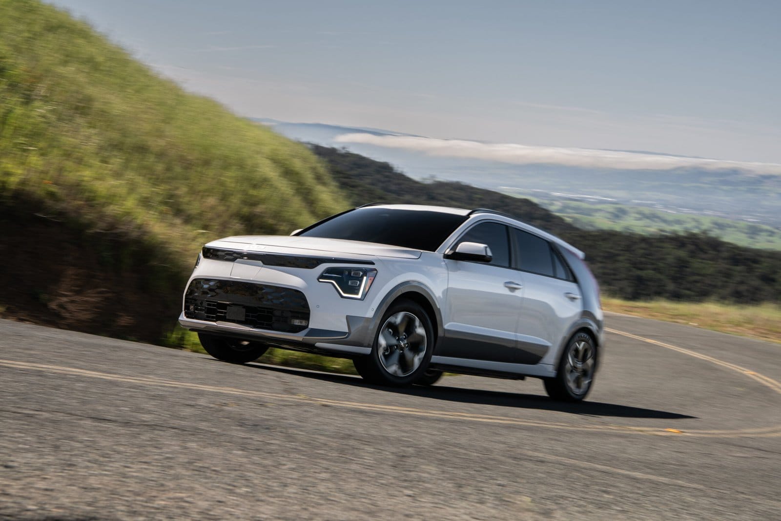 White Kia Niro EV electric vehicle driving on a mountain road with hills and clouds in the background