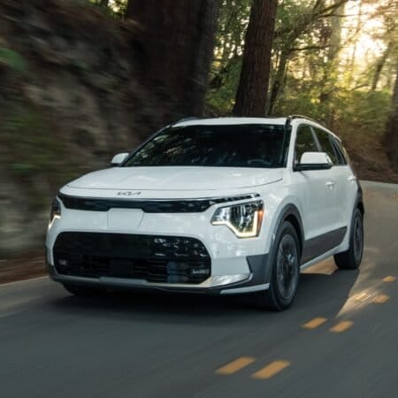 Front view of a white Kia Niro EV electric vehicle driving on a winding forest road with motion blur